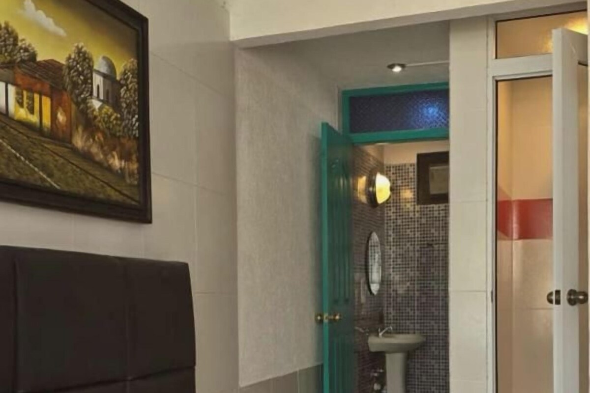 A hotel room with a bed, a painting on the wall, a bathroom with a sink and mirror, and a door leading to another room.