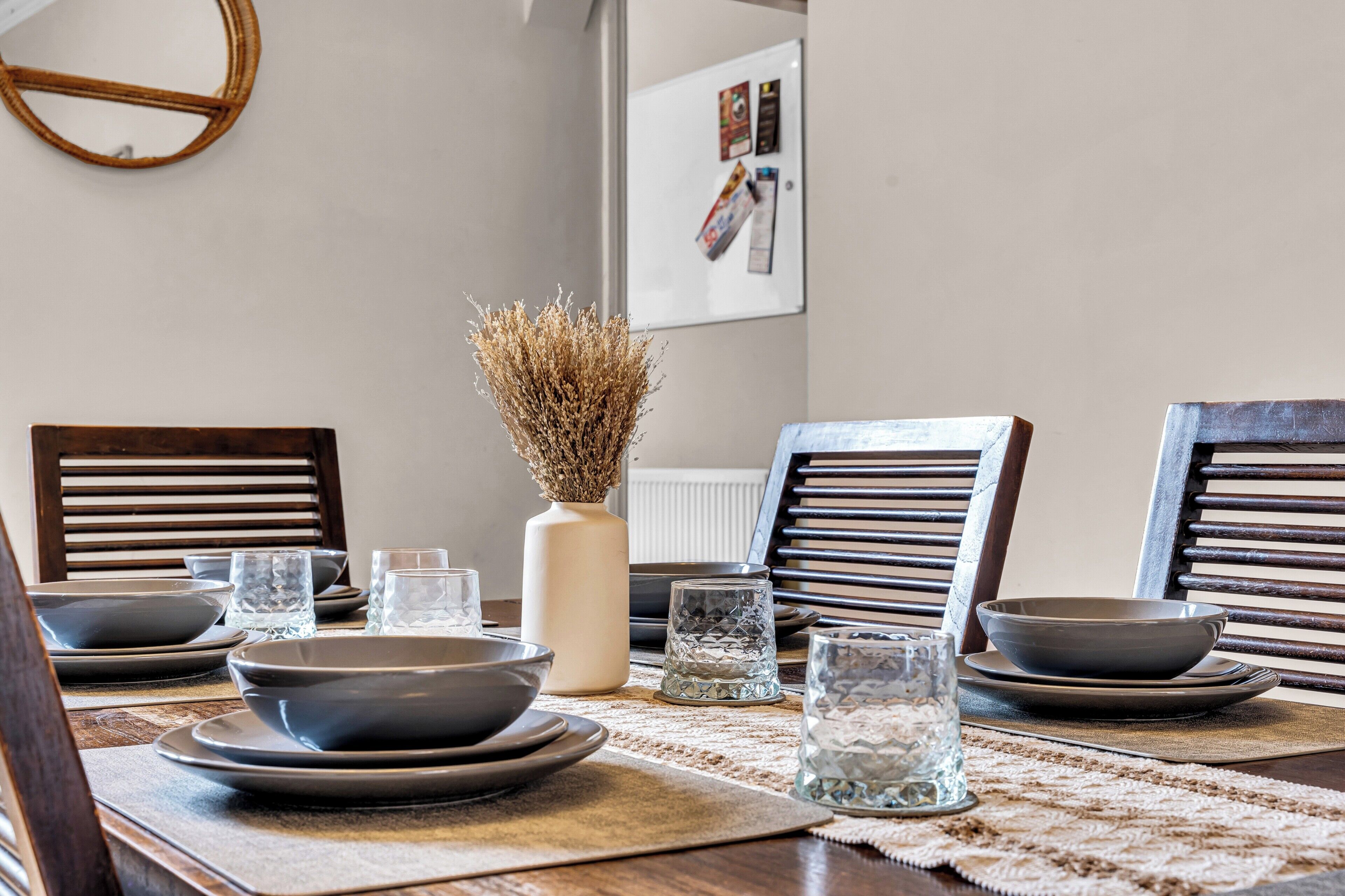 A dining table set with plates, bowls, and glasses, accompanied by chairs and a vase with dried flowers.