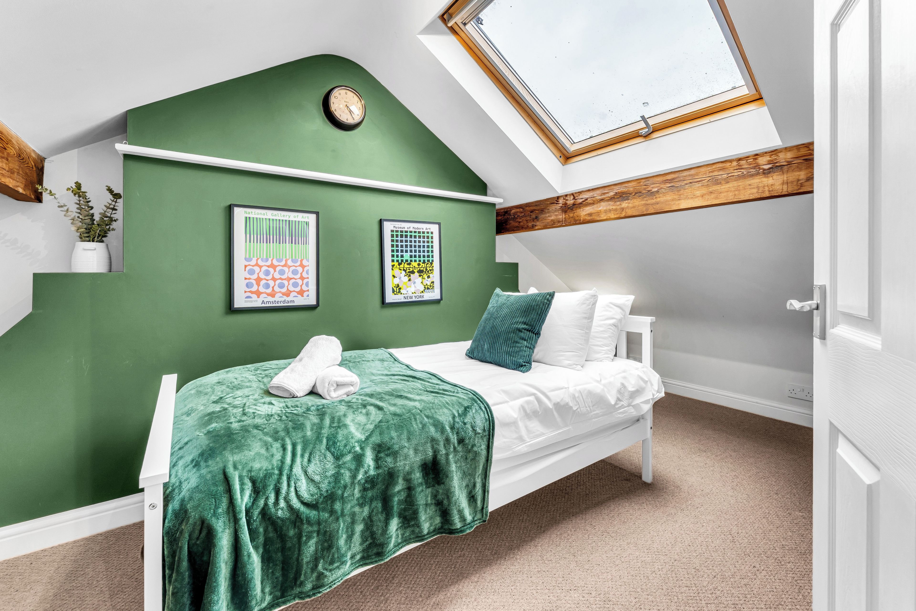 A bedroom with a sloped ceiling, a skylight, a bed with white bedding, a green wall, and two framed posters.