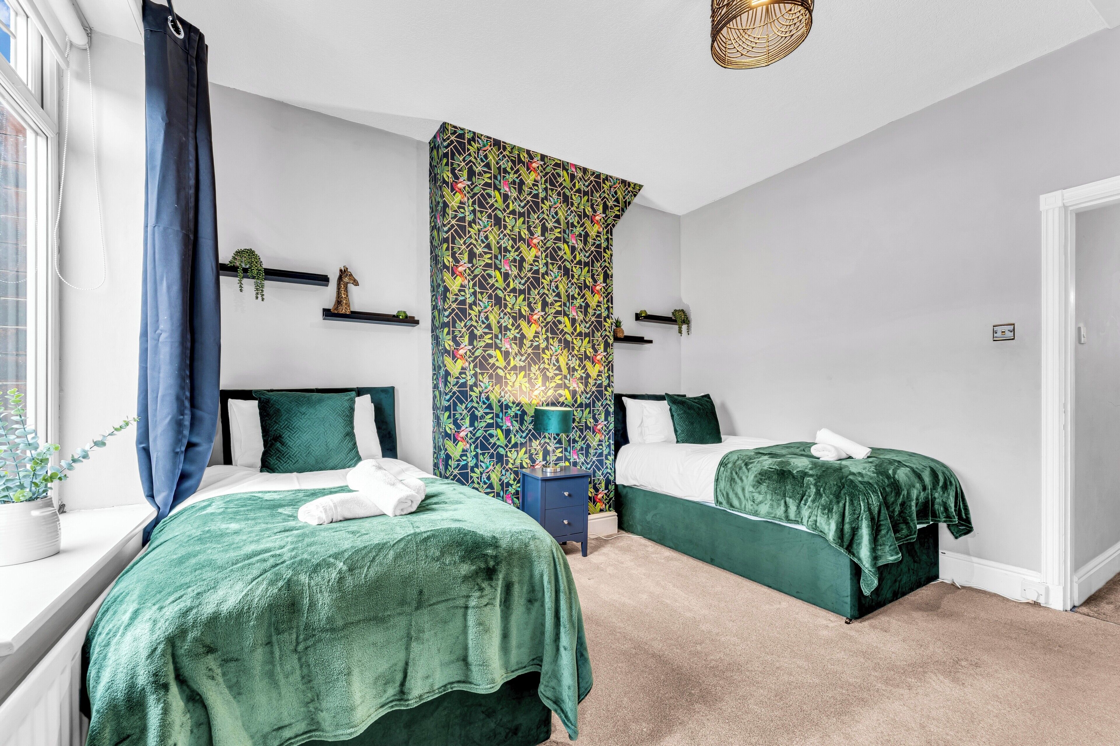 A bedroom with two beds, a window with curtains, a wall with a colorful pattern, and a small table with a lamp.