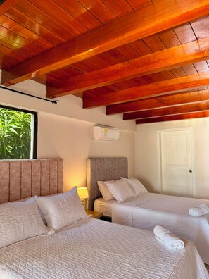A hotel room with two beds, wooden ceiling beams, a window with greenery outside, a white door, and an air conditioning unit. - Beachfront House (Magdalena)