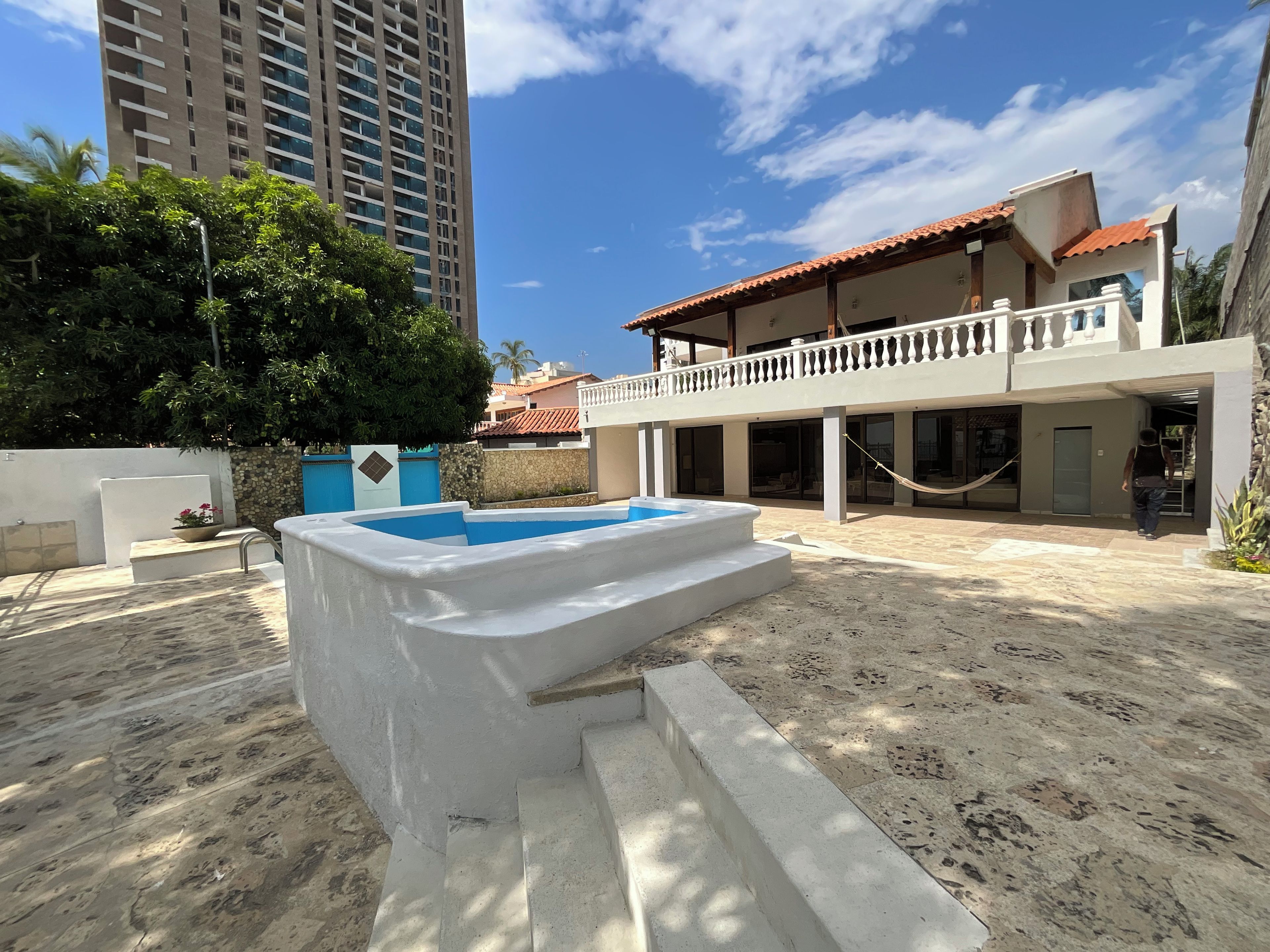 Un área de piscina con escalones de acceso, rodeada por un edificio con balcón y un edificio residencial alto al fondo.