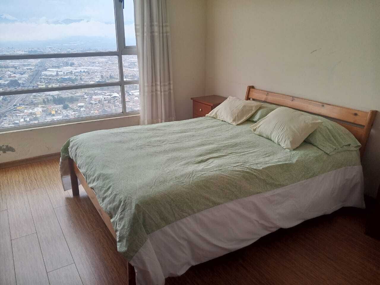 Quarto com uma janela grande, uma cama com cabeceira de madeira e uma mesinha de cabeceira.