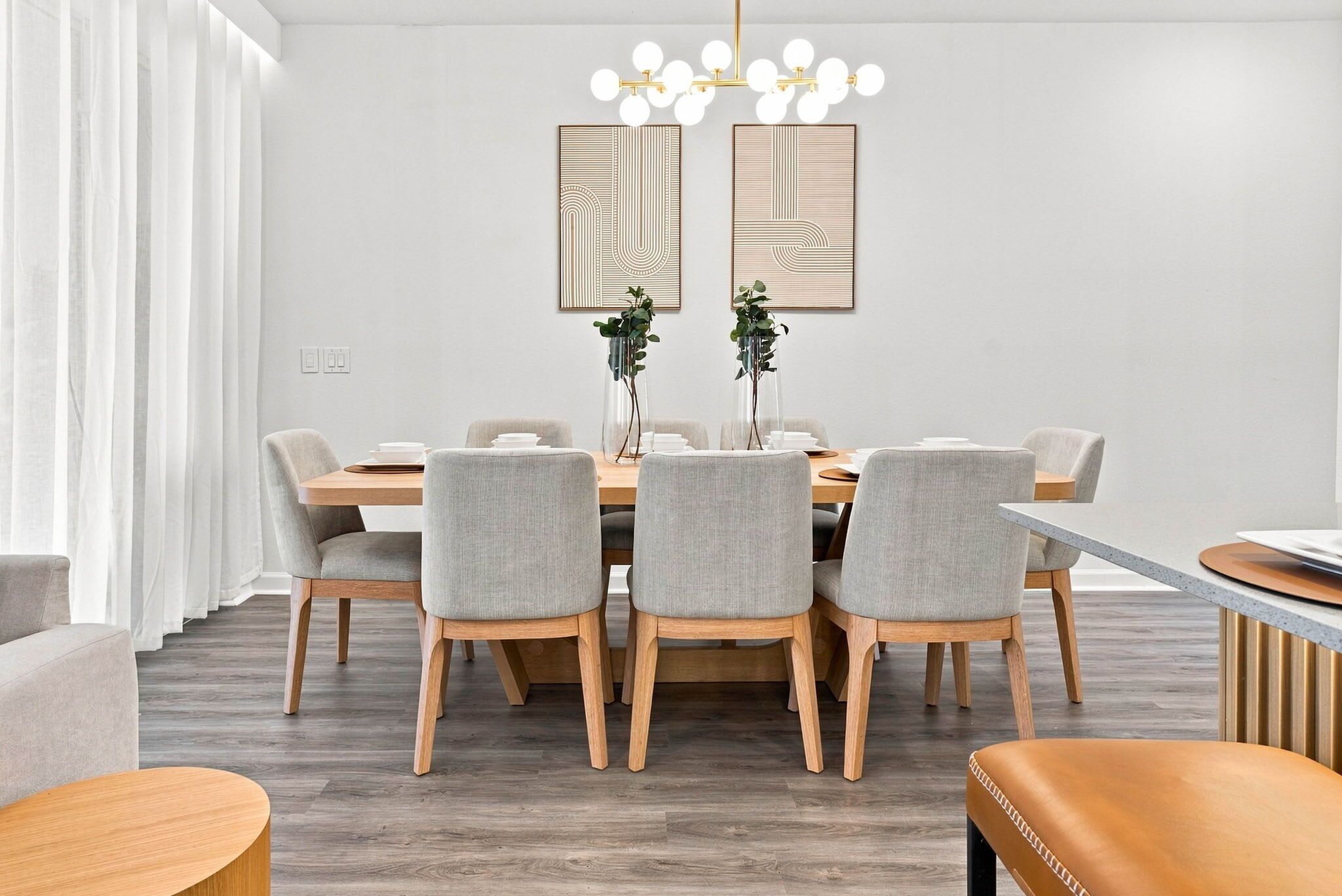 A modern dining area with a round table, six grey upholstered chairs, a wooden floor, and a chandelier with multiple light bulbs.