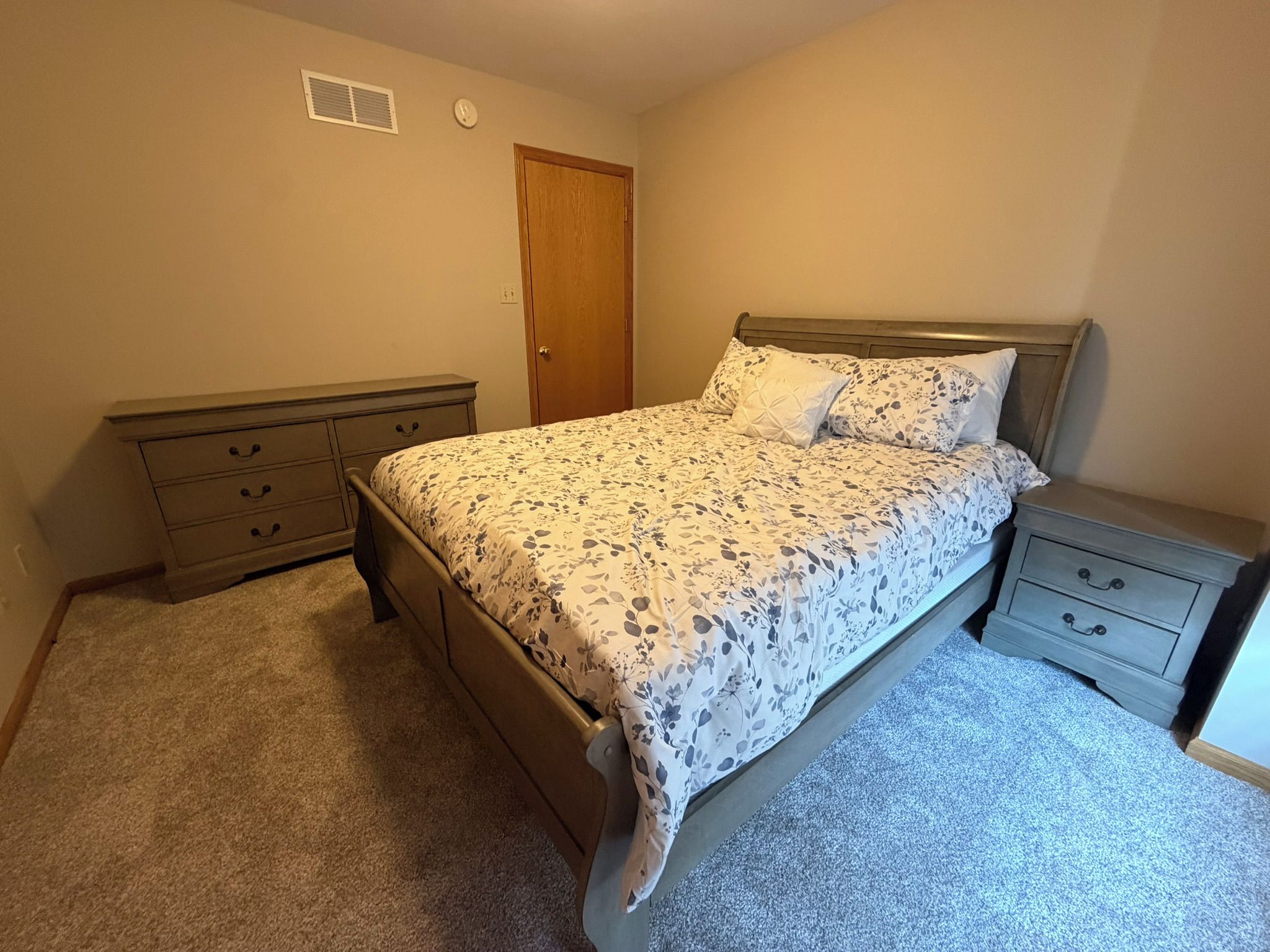 A bedroom with a bed, two nightstands, a dresser, a door, and a vent on the wall.