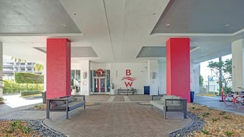 A modern building entrance with a red and white logo, two benches, and a paved area with plants.