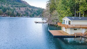 A lakeside property with a dock extending into the water, a house with a balcony, and a forested hillside in the background. - Secluded 1BR Lakefront - Dock - Deck (Port Angeles)