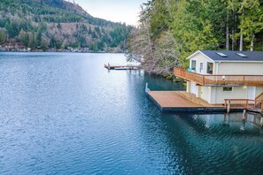 A lakeside property with a dock extending into the water, a house with a balcony, and a forested hillside in the background. - Secluded 1BR Lakefront - Dock - Deck (Port Angeles)