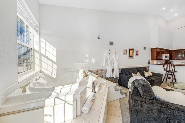 Un salon moderne avec une grande fenêtre, une baignoire intégrée, un canapé foncé et un coin cuisine à l’arrière-plan.
