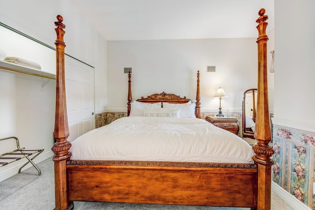 Un lit à baldaquin avec une structure en bois et du linge de lit blanc, une table de chevet avec une lampe et un miroir au mur.