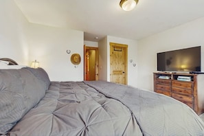 A bedroom with a large bed, a wooden wardrobe, a television, and a door leading to another room. - 2 BR Townhome at the Villages at Roslyn Ridge (Ronald)