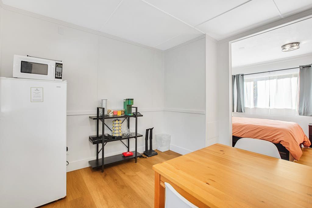 Une pièce compacte comprenant un coin cuisine, une table en bois, un lit près d’une fenêtre et une étagère contenant divers objets.