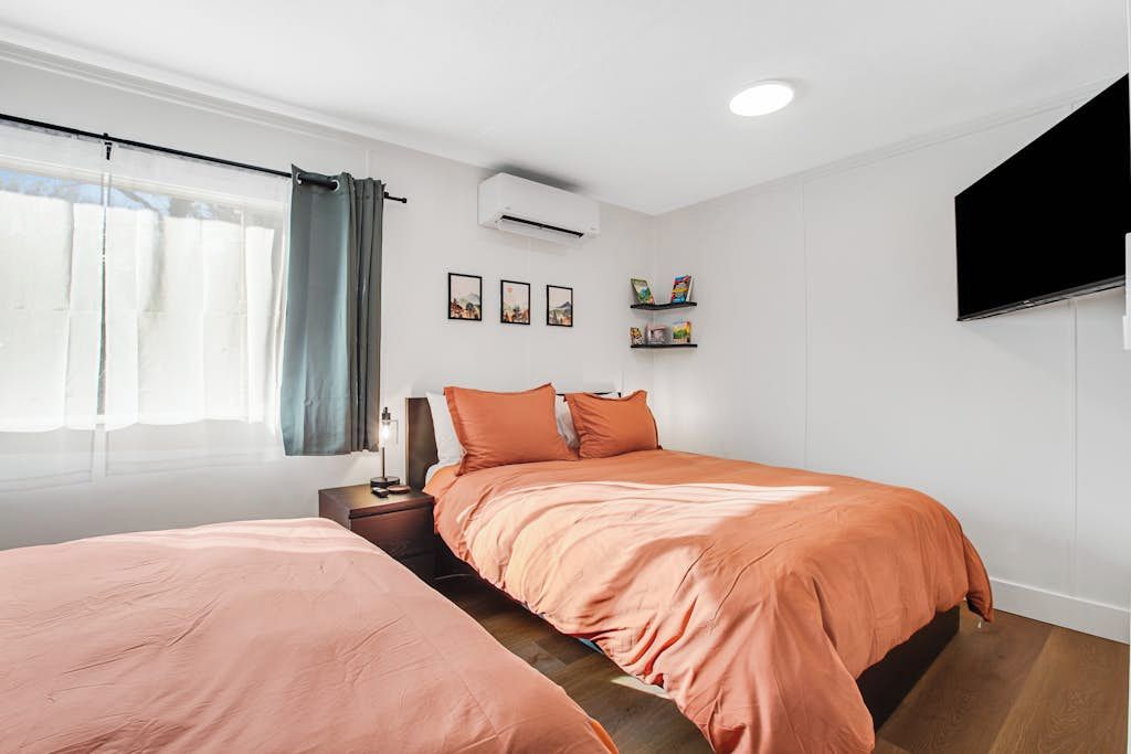A bedroom with a bed, a bedside table, a lamp, a window with curtains, and a wall-mounted TV.