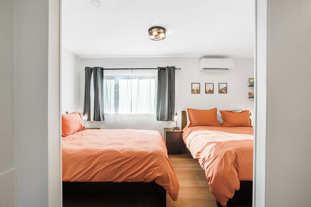 A hotel room with two beds, a window with curtains, a ceiling light, and a wall-mounted air conditioner.