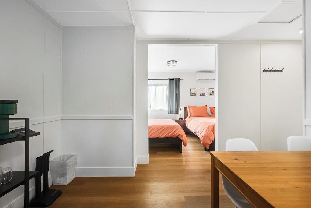 Quarto de hotel moderno com piso de madeira, cama com roupa de cama laranja, escrivaninha com cadeira e uma pequena lixeira.