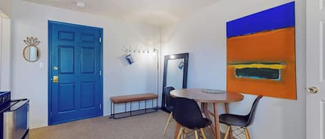 A room with a blue door, a round table with chairs, a bench, a mirror, and a large abstract painting on the wall.