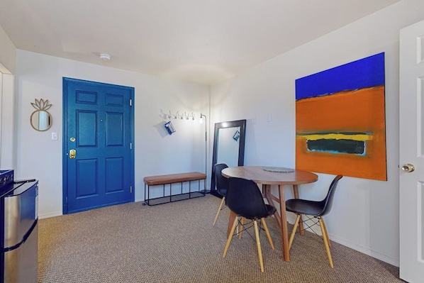 Een kamer met een blauwe deur, een ronde tafel met stoelen, een bankje, een spiegel en een groot abstract schilderij aan de muur.