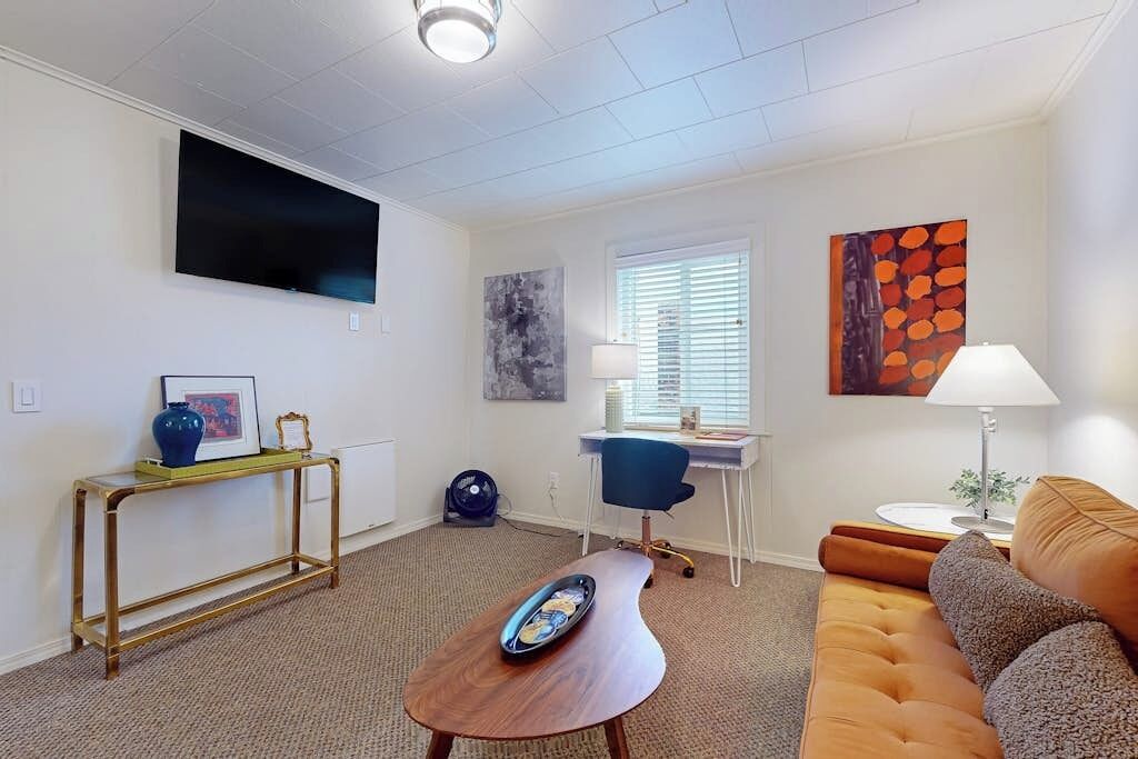 Un salon moderne doté d’un téléviseur à écran plat, d’un bureau avec une chaise, d’un canapé et d’œuvres d’art accrochées aux murs.