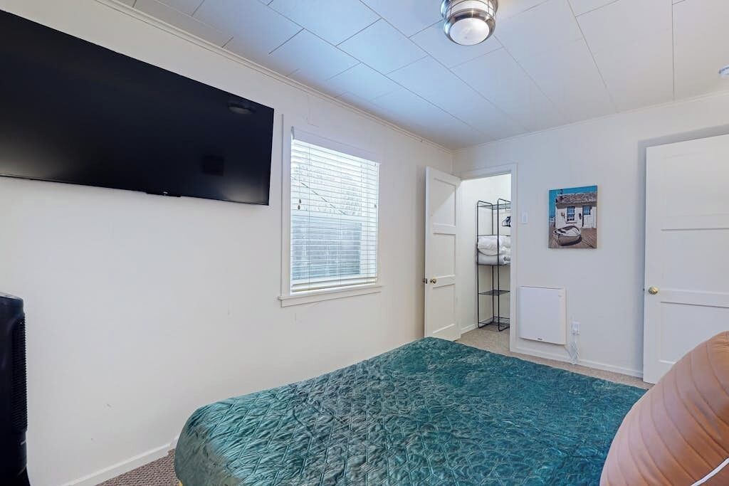 Une chambre à coucher comprenant un lit, un téléviseur fixé au mur, une fenêtre avec des stores et une porte donnant sur une salle de bain.