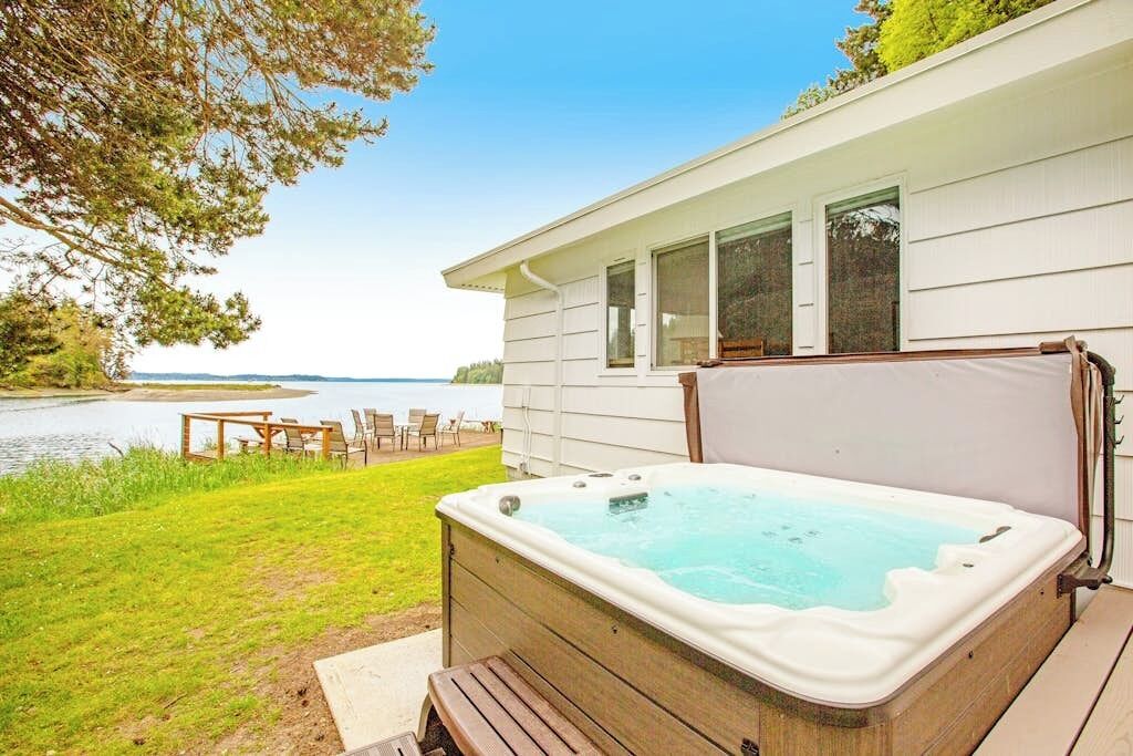 A hot tub on a wooden deck with a view of a lake and a house in the background.