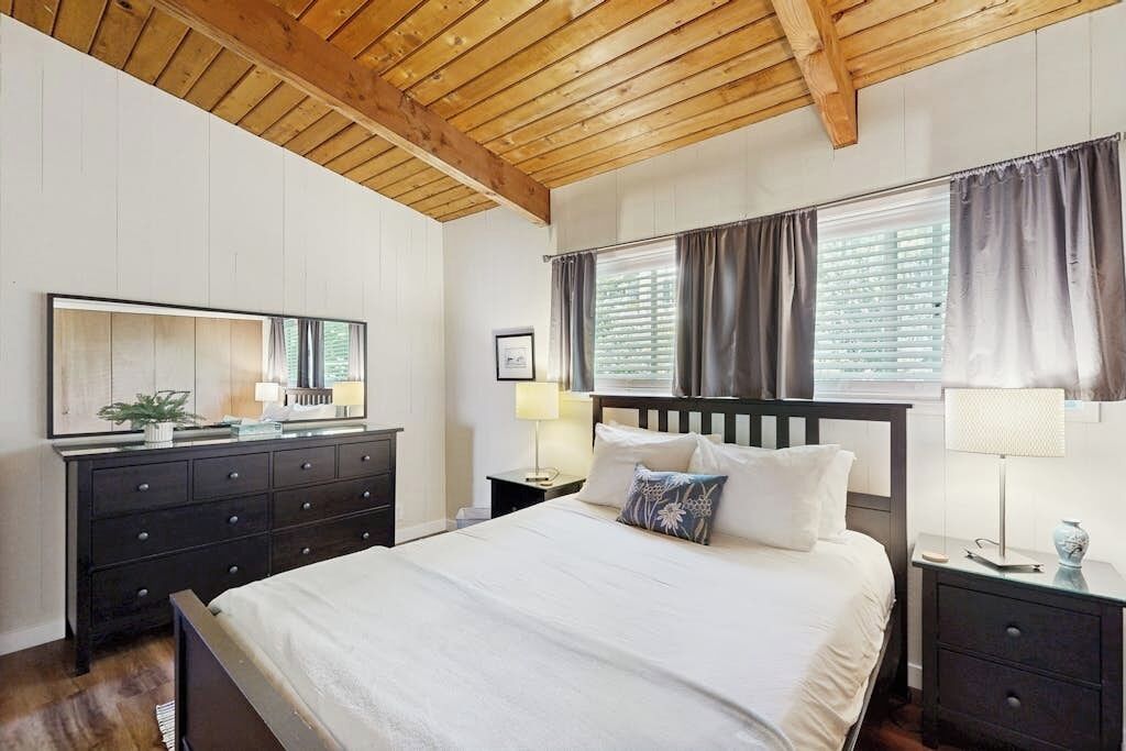 A bedroom with a wooden ceiling, a bed with white bedding, a dresser, a mirror, and two bedside lamps.