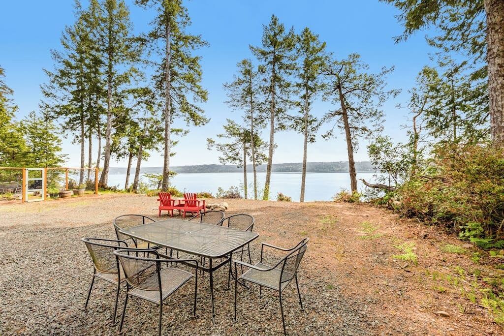 Un espace extérieur aménagé avec une table et des chaises, entouré d’arbres et offrant une vue sur une étendue d’eau.