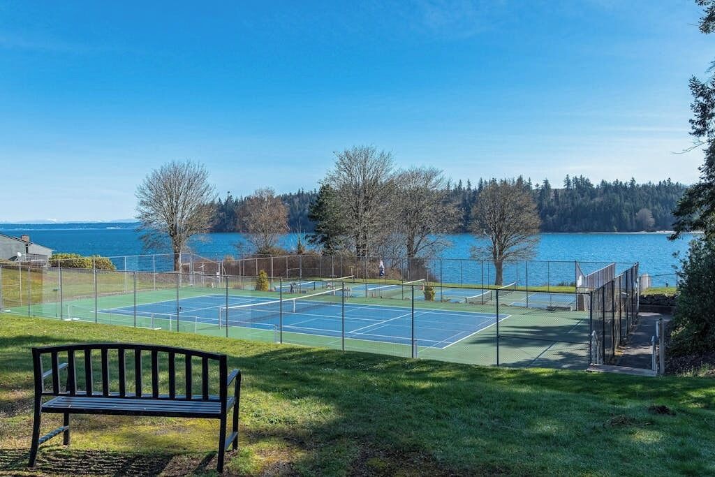Ein Tennisplatz mit Bank, umzäunt und mit Blick auf ein Gewässer und Bäume.