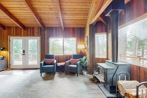 A cozy cabin interior with a fireplace, wooden walls, and a view through large windows. - 3BR Lakefront Gas Grill - Deck - Firepit (Hoodsport)