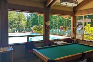 Ein Billardtisch in einem Raum mit großen Fenstern mit Blick auf einen Pool und einen Garten.