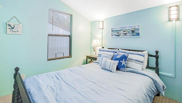 Un dormitorio con una cama, una mesita de noche, una lámpara de pared, una ventana con persianas y un cuadro.