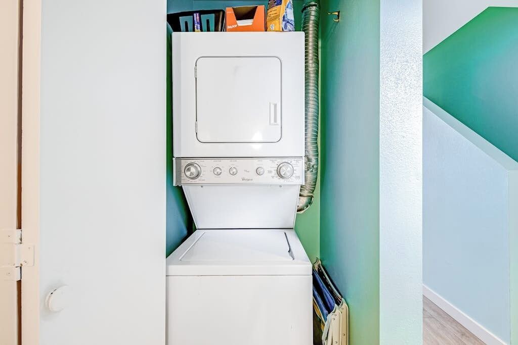 A compact laundry room with a stacked washer and dryer, a hose, and a door leading to another room.