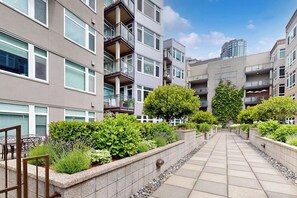 A multi-story residential building with balconies, a paved walkway, and landscaped areas with shrubs and trees. - Waterfront 1BR with fitness center & hot tub (Seattle)