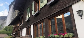Uma chalé tradicional com telhado inclinado, venezianas de madeira e montanhas ao fundo.