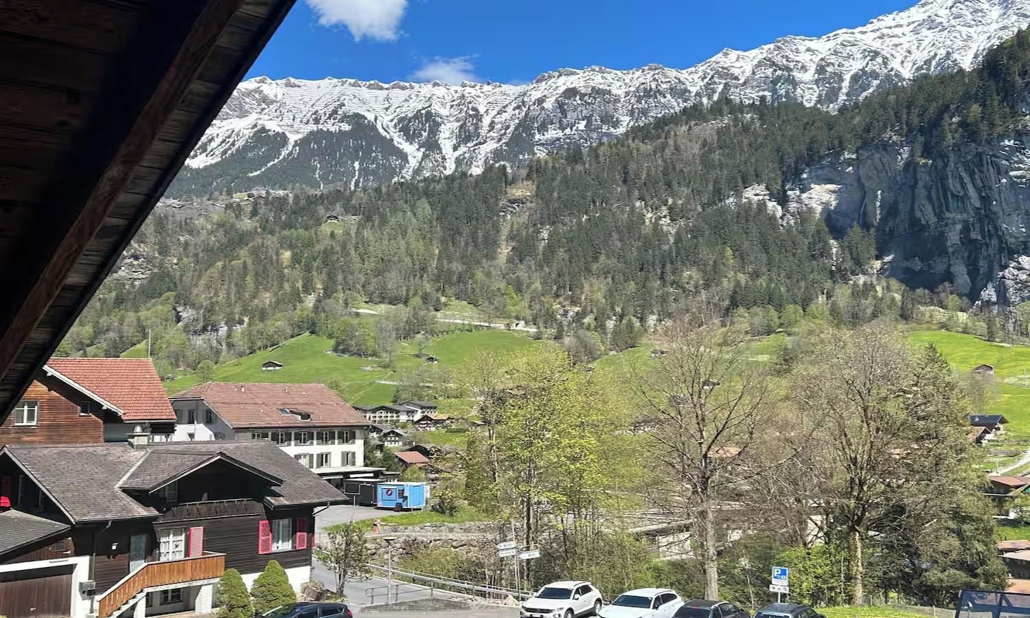 A mountain village with traditional buildings, a parking area, and a view of snow-capped mountains.