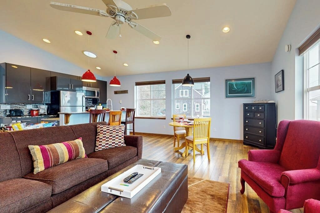 Una sala moderna con un sofá, un sillón rojo, una zona de comedor con una mesa y sillas, y una cocina al fondo.