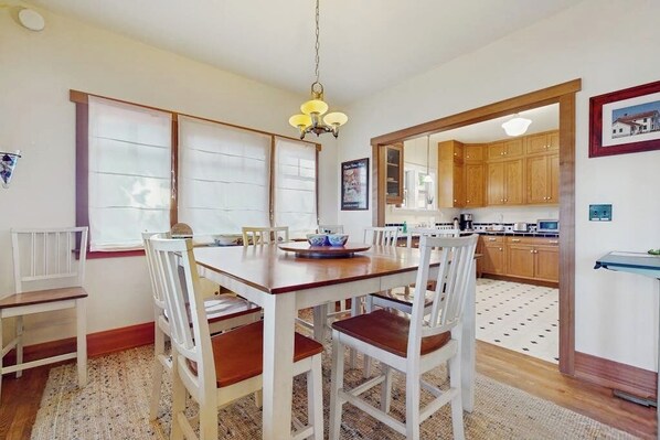 A dining area with a wooden table and white chairs, a hanging light fixture, and a kitchen in the background.