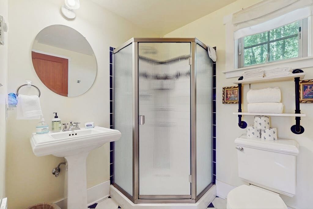 A bathroom with a glass-enclosed shower, a sink with toiletries, a toilet, and a window with a view of trees.