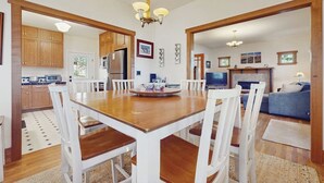 A dining area with a wooden table and white chairs, a kitchen with wooden cabinets, and a living area with a blue sofa and a television. - 3BR Oceanfront Dog Friendly | Fireplace (Port Townsend)