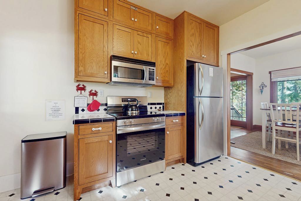 A kitchen with wooden cabinets, a stainless steel refrigerator, a microwave, an oven, and a trash bin.
