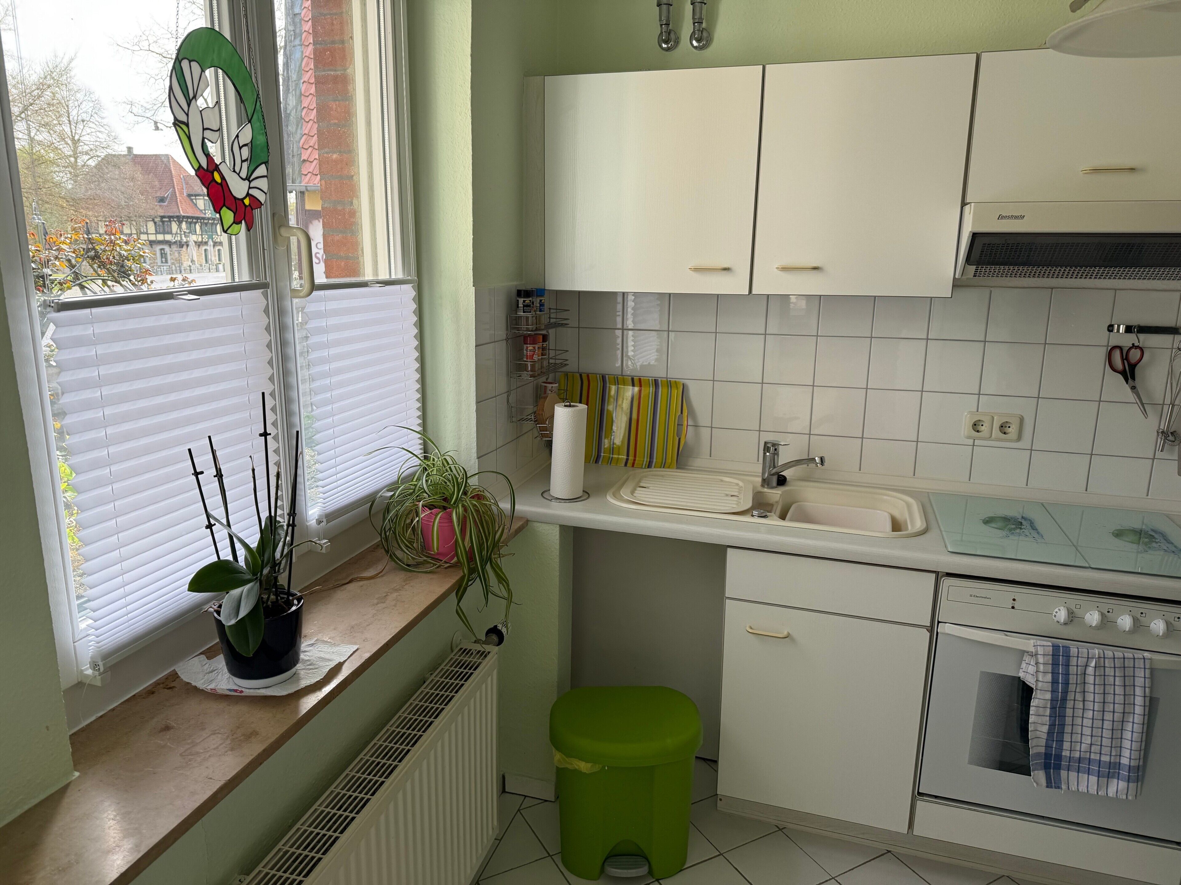 Une cuisine avec des meubles blancs, un évier et un four. Il y a une fenêtre avec des stores et une plante en pot sur le comptoir.