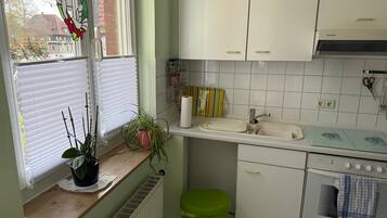 Une cuisine avec des meubles blancs, un évier et un four. Il y a une fenêtre avec des stores et une plante en pot sur le comptoir.