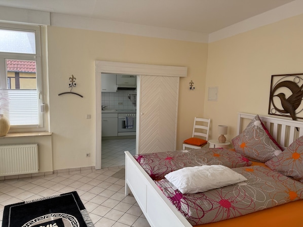 A bedroom with a bed, a chair, a table, and a view of a kitchen.