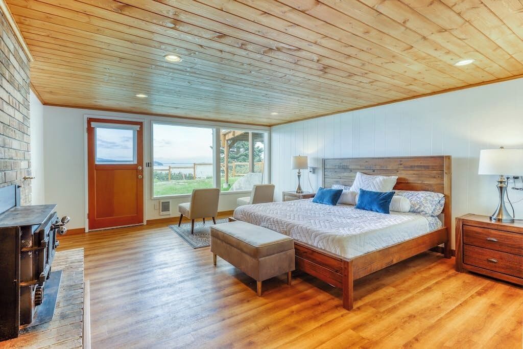 A bedroom with a wooden ceiling, a large bed, a wooden nightstand, and a view through a glass door.
