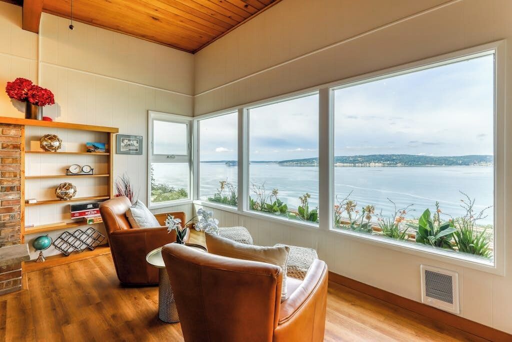 Ein Zimmer mit großem Fenster und Blick auf ein Gewässer, einer Ledersofa, einem Bücherregal und einer Holzbalkendecke.