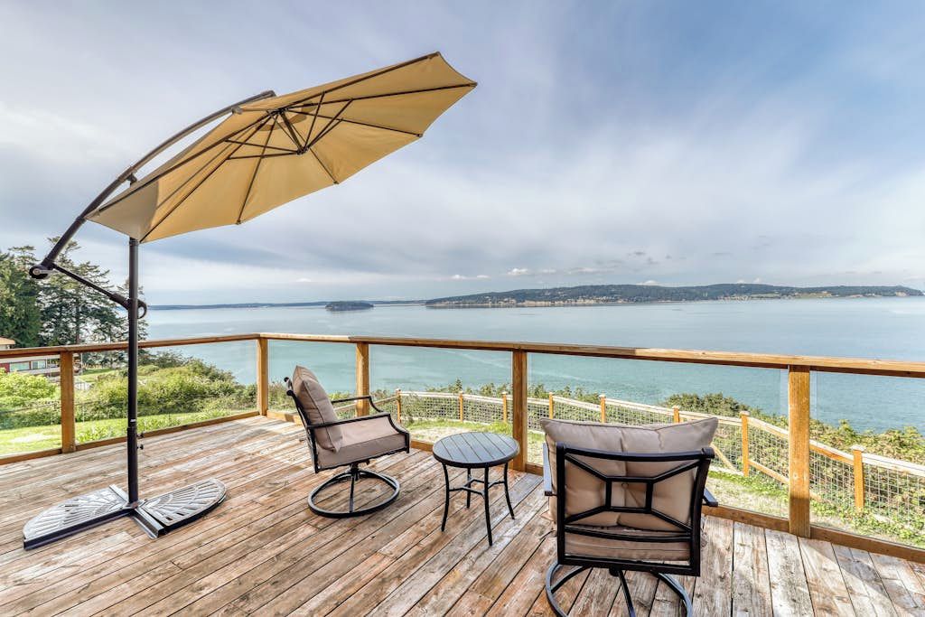 A wooden deck with outdoor furniture, including two chairs and a table, overlooking a body of water with a distant landmass.