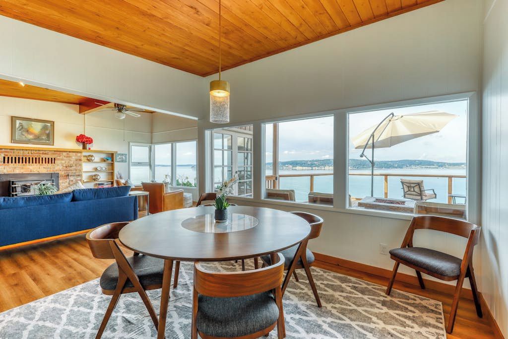A modern dining area with a round table, chairs, and a view of the sea through large windows.