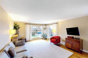 Ein Wohnzimmer mit einer Couch, einem roten Sessel, einem Fernseher und einem großen Fenster mit Vorhängen.