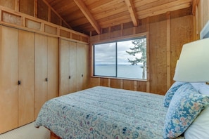 Quarto com uma janela ampla, paredes de madeira, cama com colcha estampada e mesa de cabeceira com abajur.