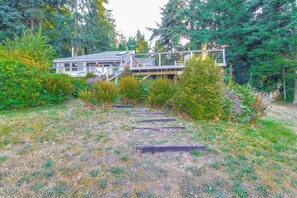 A house with a deck and a staircase leading to it, surrounded by dense greenery. - 1BR water-view cabin with fireplace (Port Ludlow)