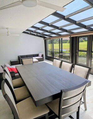 Een moderne eethoek buiten met een grote tafel en stoelen, een plafondventilator en uitzicht op een groen landschap door grote ramen.